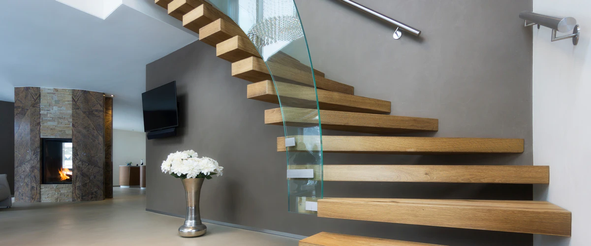 A contemporary floating stair featuring thick wooden treads extending from a grey accent wall, complemented by a seamless curved glass railing.