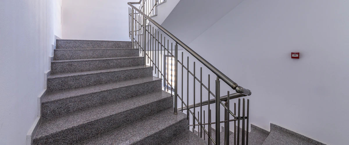 A contemporary metal stair railing with sleek vertical balusters and a cylindrical handrail installed on a grey speckled granite staircase.