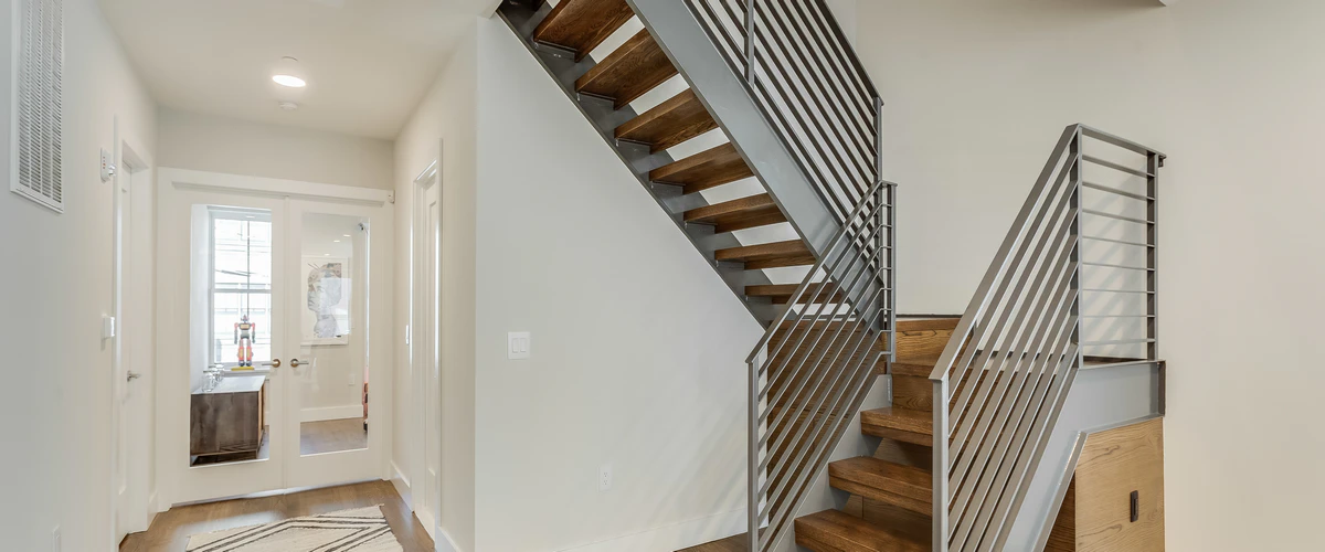 Custom contemporary metal stair railing featuring horizontal and vertical bars on a wooden staircase in a bright hallway.