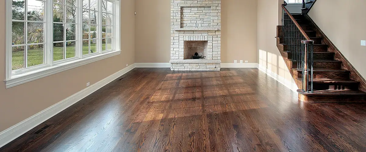 Dark brown hardwood floor in a spacious living room featuring a stone fireplace, large bright windows, and a wooden staircase.