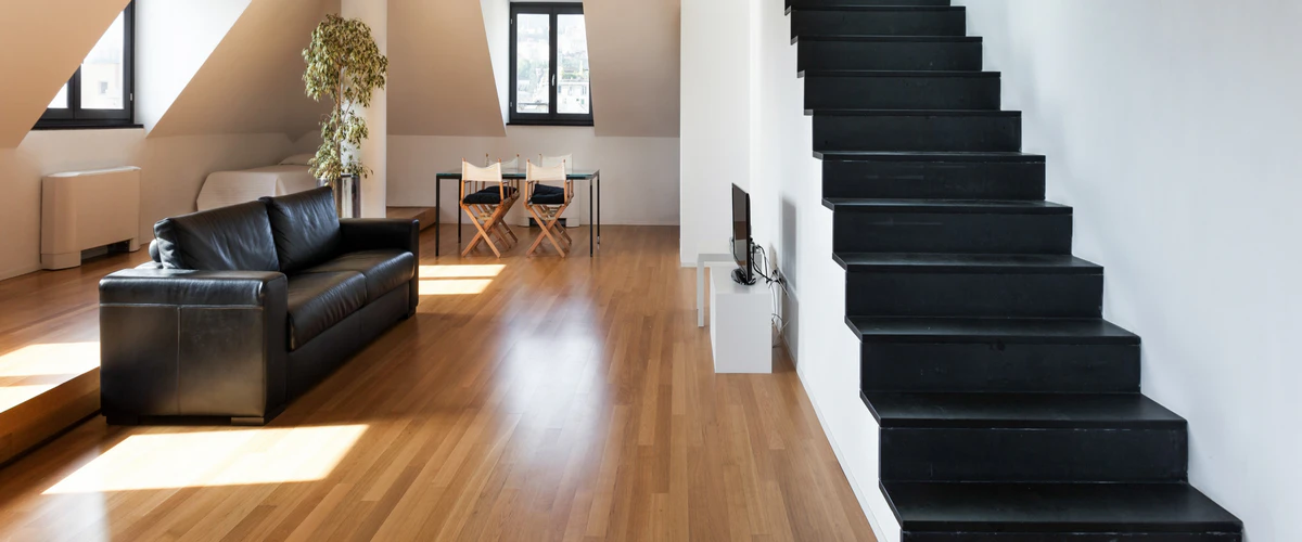 A minimalist modern living space anchored by a seamless medium-toned hardwood floor, featuring a black leather sofa and contrasting black open-tread stairs.