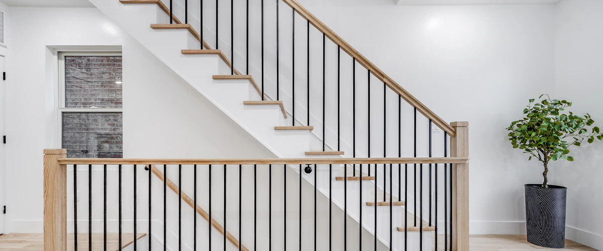 Stair Railing Installation in Alexandria, VA 2 Side view of a modern minimalist interior staircase with wood treads and white stringers, highlighting a sleek railing design made of straight black metal balusters and a light wood handrail, situated next to a potted plant.