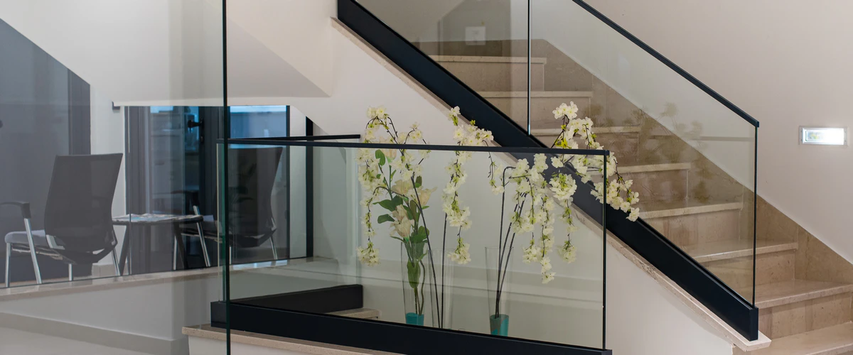 A modern commercial stair design featuring light stone steps and a transparent glass balustrade with a black handrail, accented by white floral arrangements on the landing.