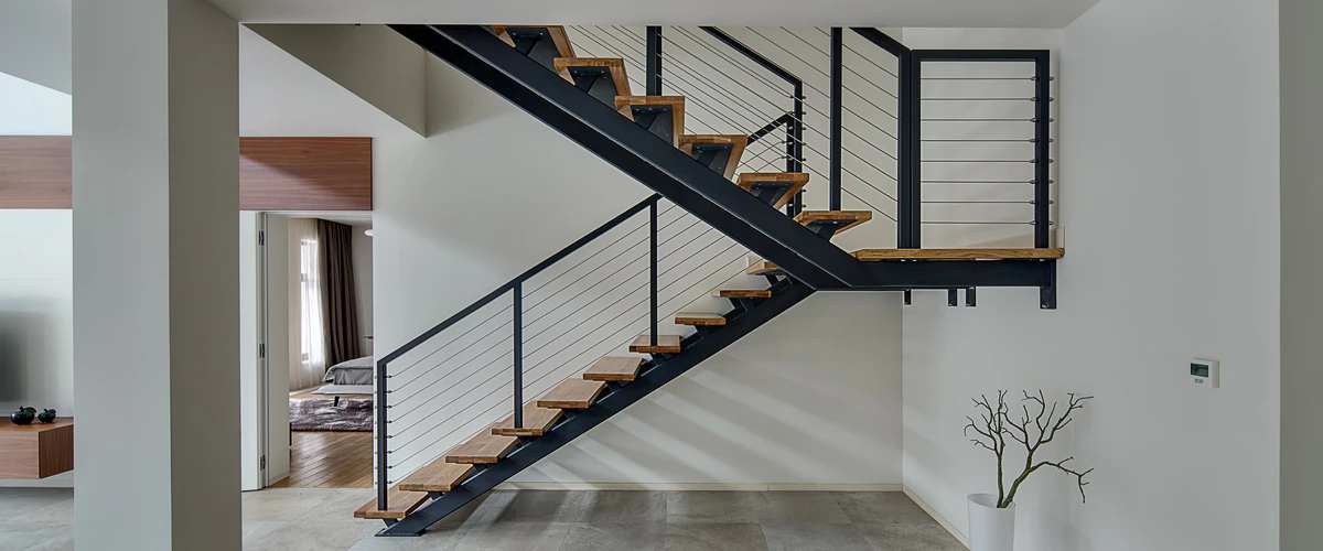 Modern minimalist stair featuring light wood treads supported by black metal stringers and a horizontal wire cable railing in a bright white residential interior.