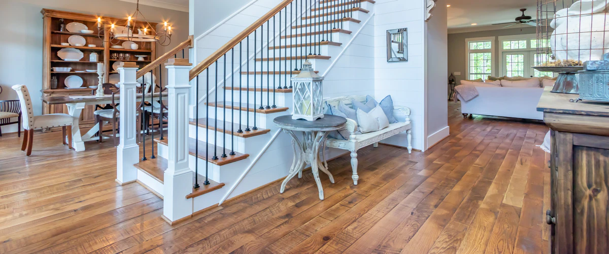 A bright, open-concept dining and living area featuring a wide-plank rustic hardwood floor, a large wooden hutch, and a white staircase with wooden treads.